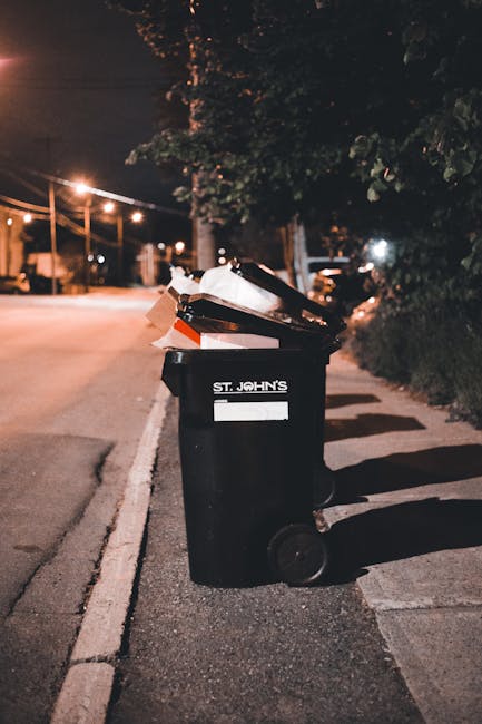 A black plastic wheelie bin with a white label and the words 'ST. JOHN'S' printed on the front, positioned on a residential street sidewalk during nighttime. The bin is open and overflowing with vario